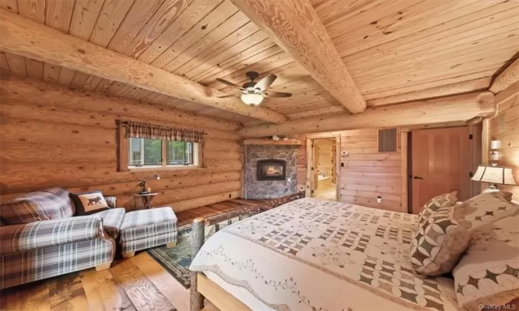 Bedroom with log walls, beamed ceiling, hardwood / wood-style floors, wooden ceiling, and a stone fireplace