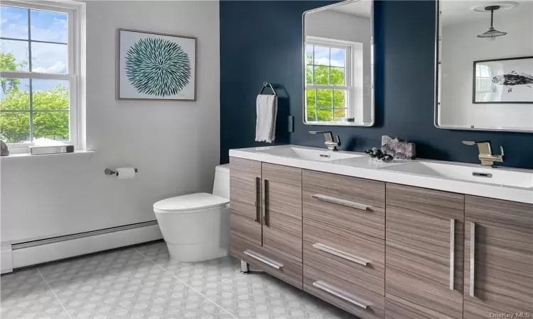 Bathroom featuring dual bowl vanity, tile flooring, baseboard heating, and toilet