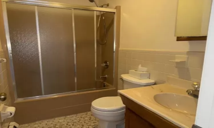 Full bathroom with tile walls, vanity, combined bath / shower with glass door, and toilet