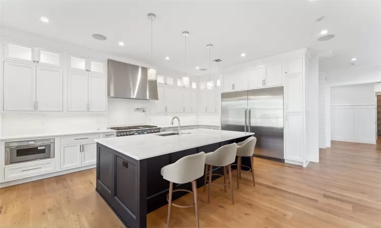 Kitchen with white cabinetry, wall chimney exhaust hood, stainless steel appliances, light hardwood / wood-style floors, and a kitchen island with sink