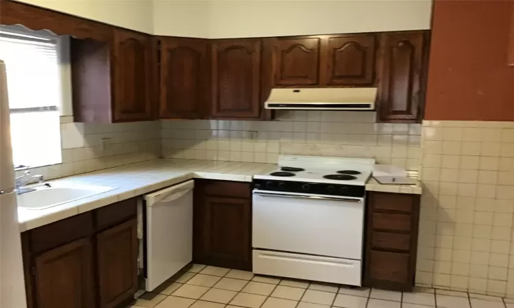 Kitchen with white appliances, extractor fan, sink, light tile patterned floors, and tile counters