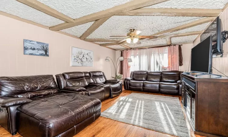 Living room featuring a textured ceiling, ceiling fan, wooden walls, beam ceiling, and light hardwood / wood-style flooring