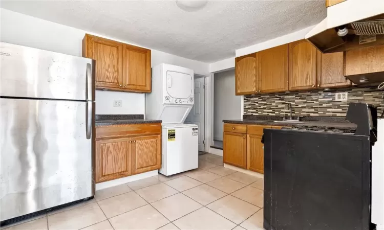 Kitchen featuring stacked washer and clothes dryer, backsplash, ventilation hood, light tile patterned flooring, and stainless steel refrigerator