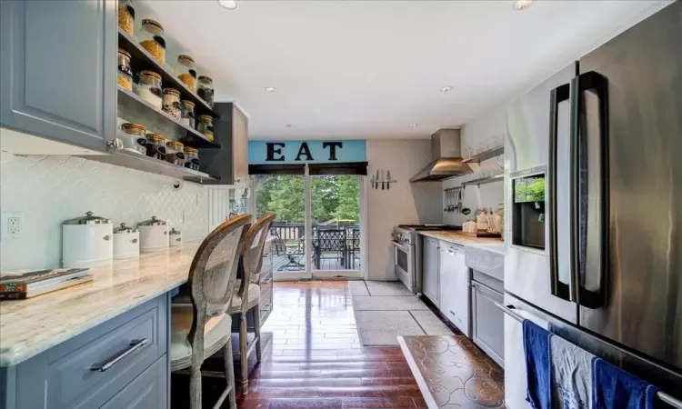 Kitchen with tasteful backsplash, wall chimney range hood, stainless steel appliances, light wood-type flooring, and light stone counters