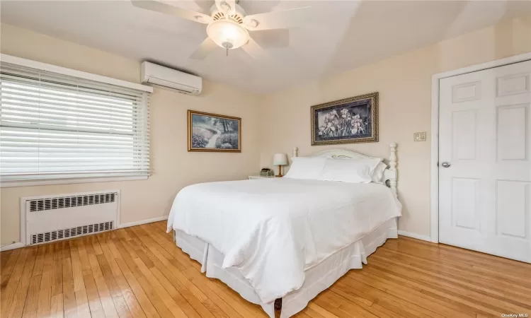 Bedroom with a wall mounted air conditioner, ceiling fan, radiator heating unit, and light hardwood / wood-style flooring