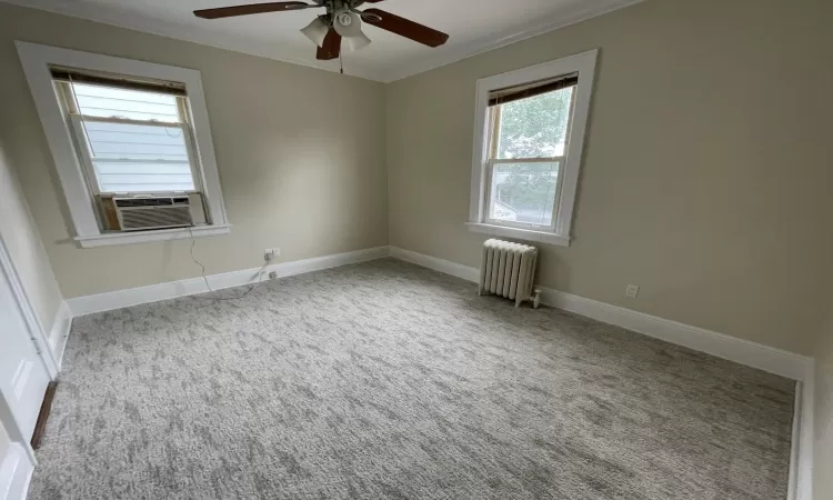 Carpeted empty room with ceiling fan, cooling unit, radiator heating unit, and crown molding