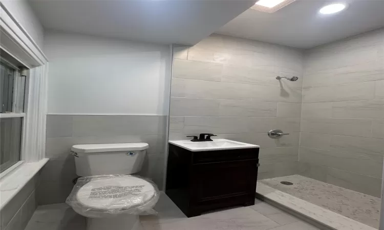 Bathroom featuring a tile shower, vanity, tile walls, and toilet