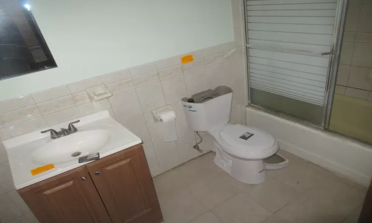 Full bathroom featuring vanity, bath / shower combo with glass door, tile patterned flooring, toilet, and tile walls