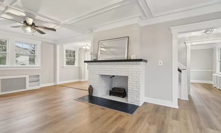 Unfurnished living room featuring a brick fireplace, radiator, ornamental molding, coffered ceiling, and ceiling fan