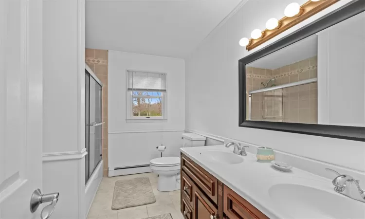 Full bathroom with vanity, a baseboard heating unit, shower / bath combination with glass door, tile patterned flooring, and toilet