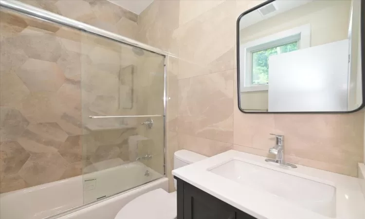 Full bathroom with backsplash, bath / shower combo with glass door, toilet, vanity, and tile walls