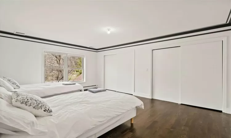 Bedroom with ornamental molding, dark wood-type flooring, a baseboard radiator, and two closets