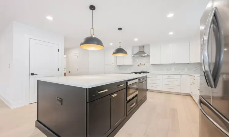 Kitchen with appliances with stainless steel finishes, wall chimney range hood, decorative light fixtures, white cabinets, and a kitchen island