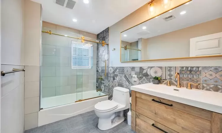 Full bathroom with tile patterned floors, bath / shower combo with glass door, toilet, decorative backsplash, and vanity