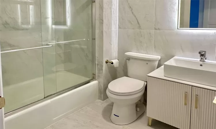 Full bathroom with vanity, toilet, bath / shower combo with glass door, and tile walls