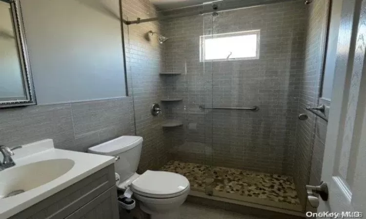 Bathroom featuring vanity, toilet, a shower with shower door, and tile walls, Stone flooring in shower.