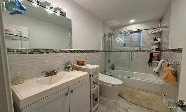 Full bathroom featuring tasteful backsplash, bath / shower combo with glass door, toilet, vanity, and tile walls