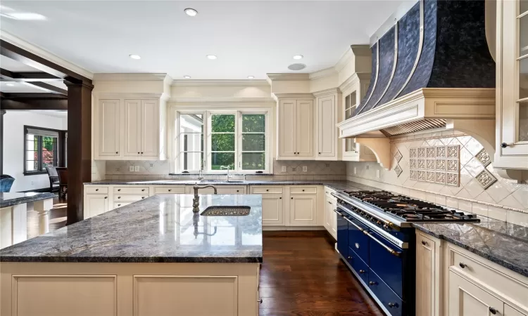 Kitchen featuring dark stone counters, cream cabinets, a center island with sink, gas stove, and custom range hood