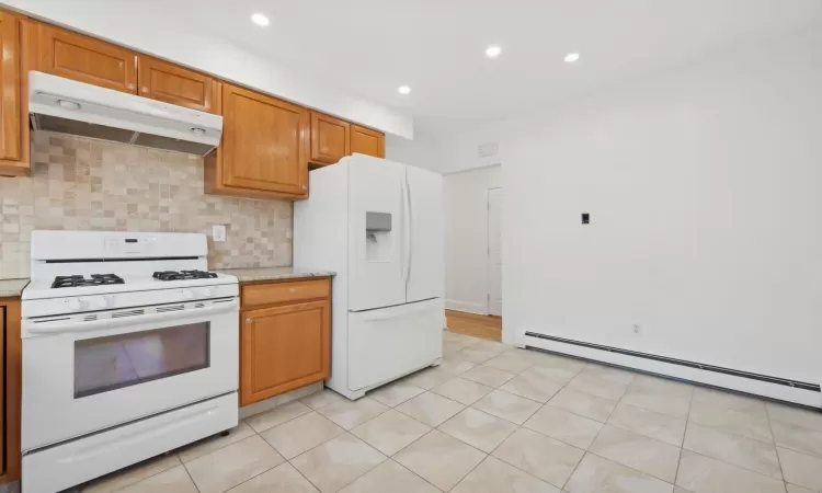 Kitchen featuring white appliances, decorative backsplash, light stone countertops, baseboard heating, and light tile patterned floors