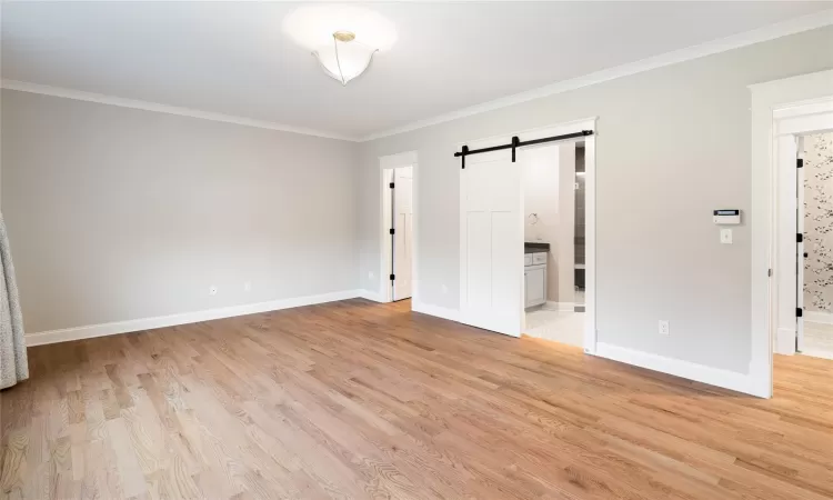 Unfurnished bedroom featuring crown molding, light wood-type flooring, ensuite bathroom, and a barn door