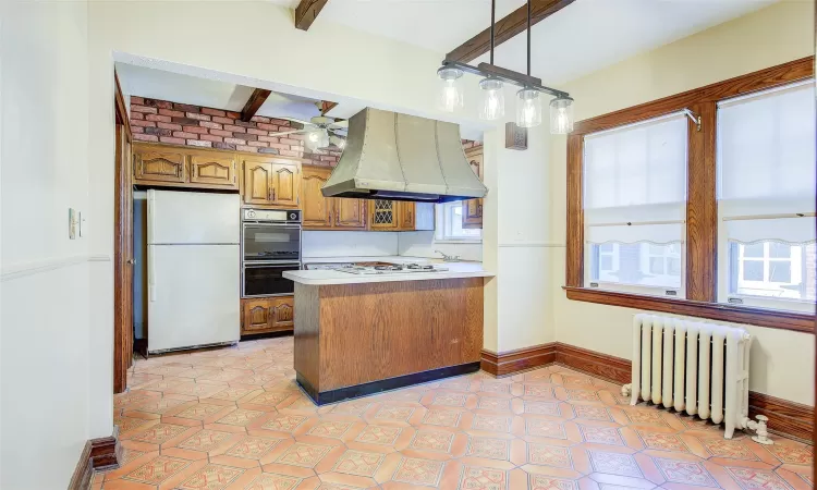 Kitchen featuring radiator heating unit, hanging light fixtures, kitchen peninsula, island exhaust hood, and white appliances