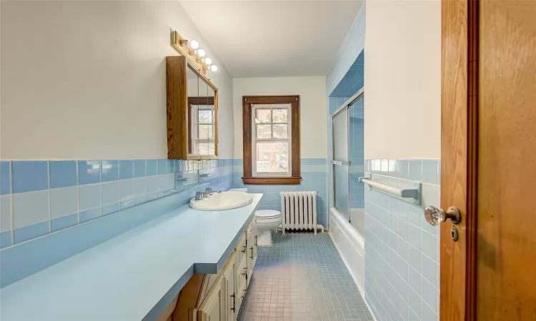 Full bathroom with combined bath / shower with glass door, vanity, tile walls, tile patterned flooring, and radiator heating unit