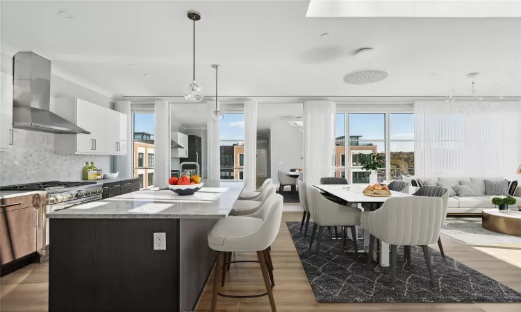 Kitchen with light stone countertops, wall chimney range hood, an island with sink, decorative light fixtures, and decorative backsplash