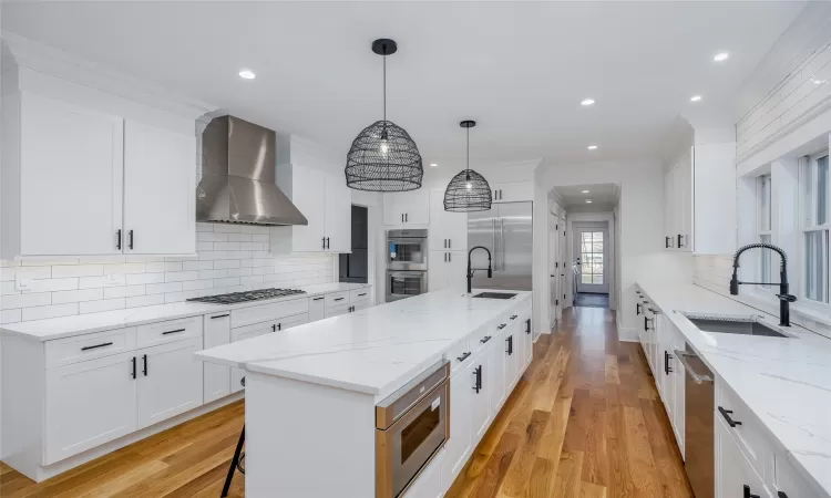 Kitchen with appliances with stainless steel finishes, wall chimney exhaust hood, decorative light fixtures, white cabinetry, and a kitchen island with sink