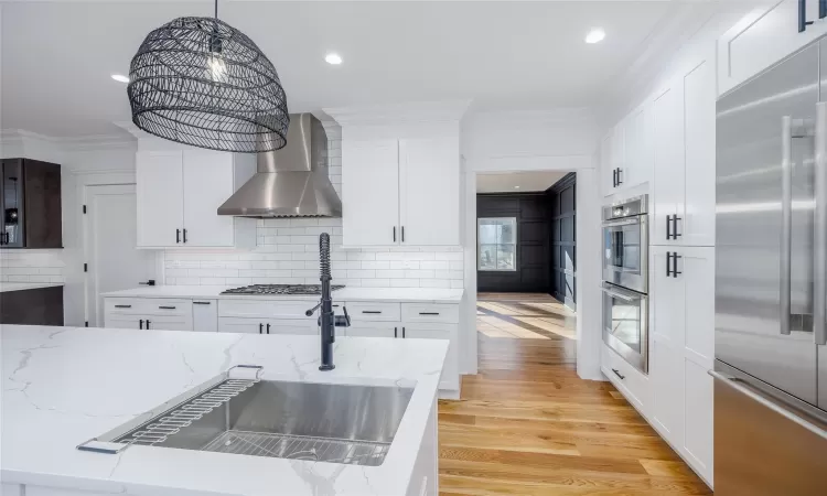 Kitchen with white cabinets, wall chimney exhaust hood, stainless steel appliances, hanging light fixtures, and light hardwood / wood-style flooring