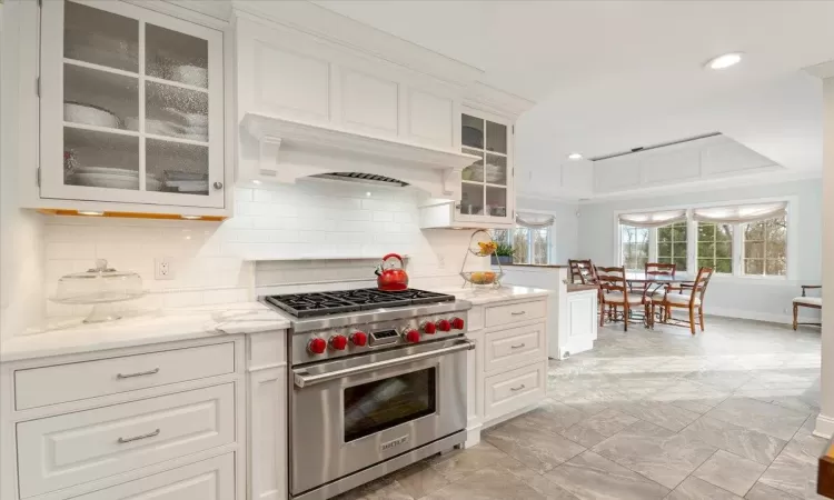 Kitchen with decorative backsplash, white cabinetry, light stone countertops, custom range hood, and luxury stove