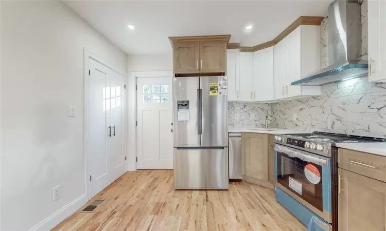 Kitchen with decorative backsplash, appliances with stainless steel finishes, wall chimney range hood, light hardwood / wood-style flooring, and white cabinets