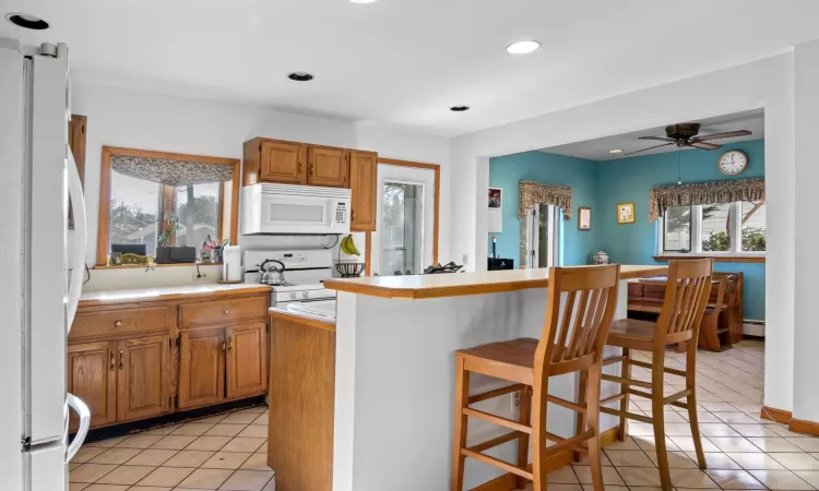 Kitchen featuring white appliances, a kitchen bar, light tile patterned flooring, ceiling fan, and a baseboard radiator