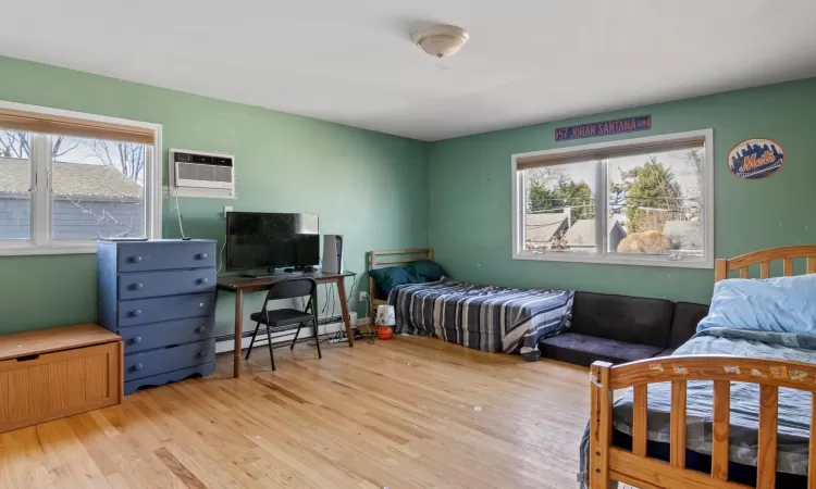 Bedroom featuring light hardwood / wood-style floors, baseboard heating, multiple windows, and a wall unit AC