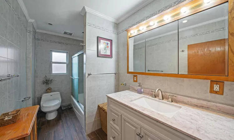 Full bathroom with toilet, enclosed tub / shower combo, tile walls, ornamental molding, and vanity