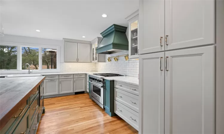 Kitchen with custom exhaust hood, backsplash, stainless steel stove, light hardwood / wood-style flooring, and light stone counters