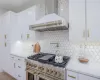 Kitchen with double oven range, white cabinetry, decorative backsplash, light stone countertops, and wall chimney range hood