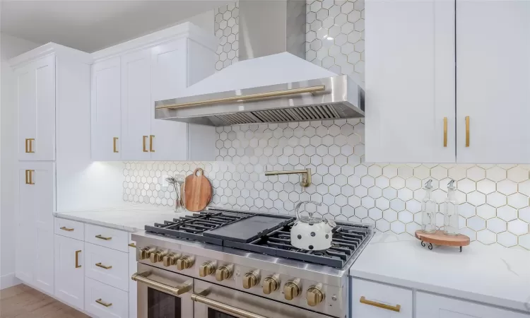 Kitchen with double oven range, white cabinetry, decorative backsplash, light stone countertops, and wall chimney range hood