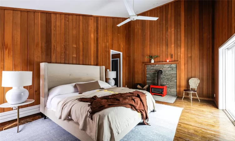 Bedroom featuring ceiling fan, a wood stove, a baseboard radiator, and wood walls