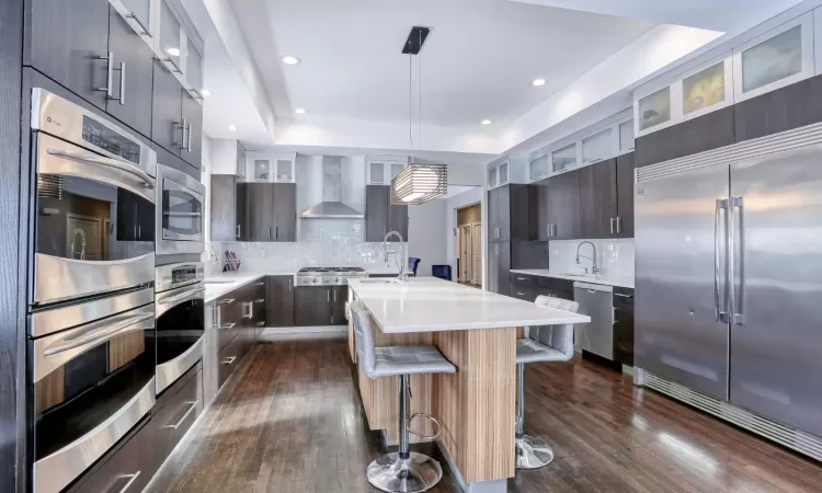 Kitchen with hanging light fixtures, a raised ceiling, stainless steel appliances, a center island with sink, and wall chimney exhaust hood