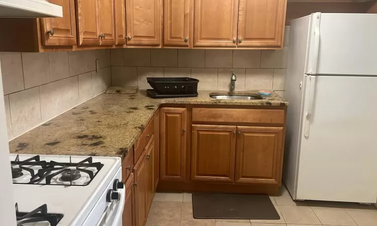 Kitchen featuring white appliances, sink, range hood, light tile patterned flooring, and light stone counters