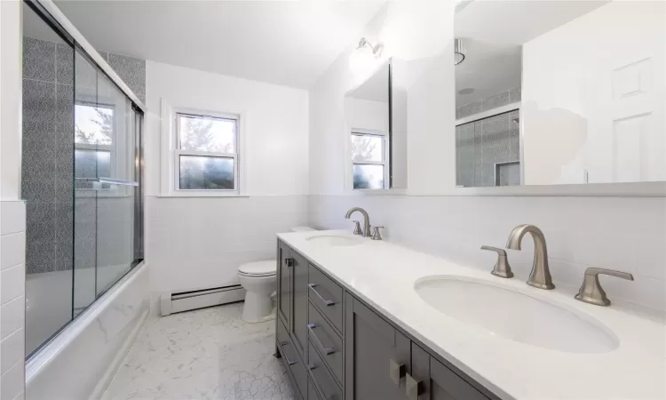 Full bathroom with baseboard heating, vanity, bath / shower combo with glass door, tile walls, and toilet