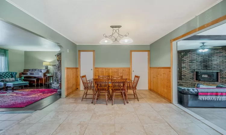 Tiled dining area featuring crown molding, ceiling fan with notable chandelier, a brick fireplace, and wood walls