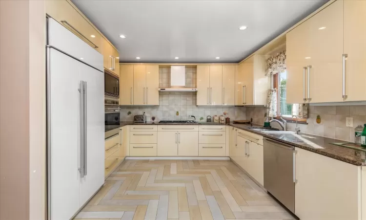 Kitchen featuring sink, built in appliances, wall chimney exhaust hood, dark stone countertops, and light parquet flooring