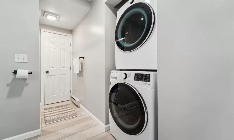 Clothes washing area featuring stacked washer / drying machine, light wood-type flooring, and a baseboard heating unit