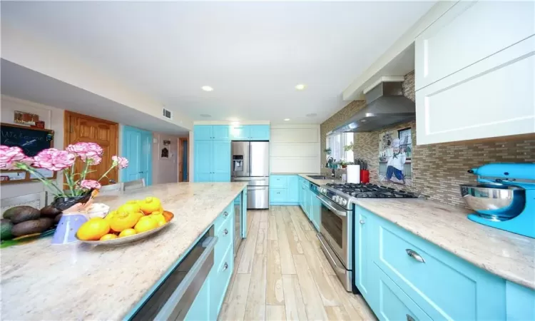 Kitchen featuring appliances with stainless steel finishes, backsplash, blue cabinetry, wall chimney range hood, and light hardwood / wood-style flooring