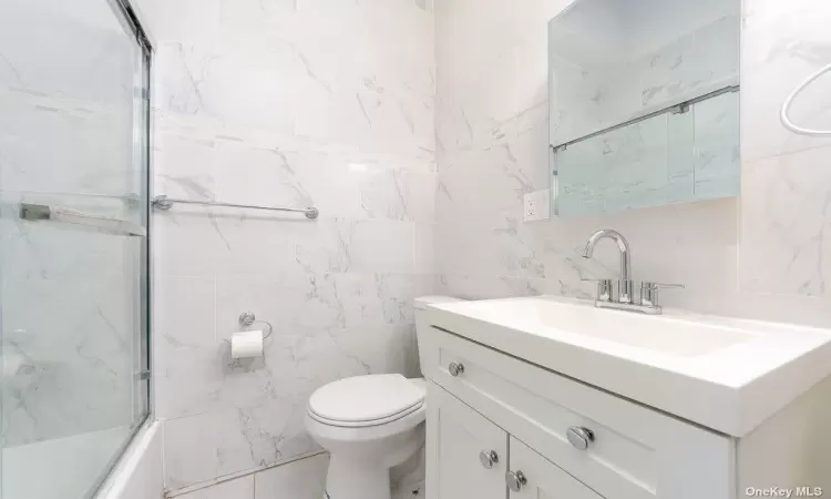 Bathroom with toilet, shower / bath combination with glass door, tile walls, and vanity