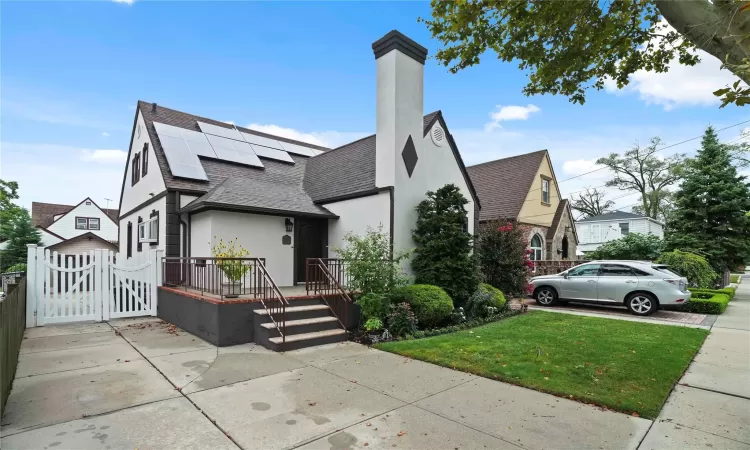 View of front of house with stucco siding, roof with shingles, a gate, roof mounted solar panels, and a front yard