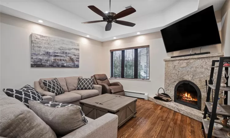 Living area featuring dark wood finished floors, ceiling fan, a stone fireplace, a baseboard heating unit, and recessed lighting