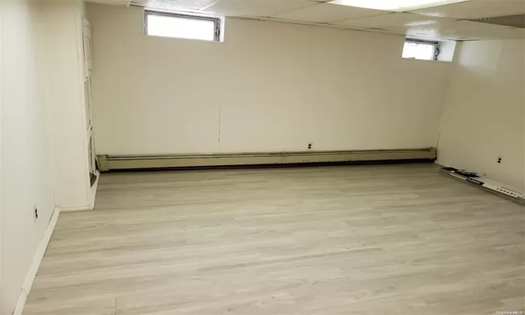 Basement with a healthy amount of sunlight, light wood-style floors, a baseboard radiator, and a drop ceiling