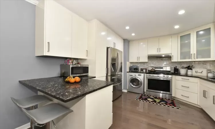 Kitchen featuring under cabinet range hood, stainless steel appliances, dark wood-type flooring, tasteful backsplash, and washer / dryer